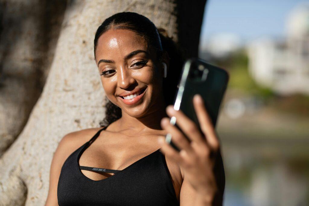 A young woman smiling as she takes a selfie outdoors, leaning against a tree in workout attire. Her bright expression and relaxed pose capture a carefree, flirty moment perfect for sharing on social media.