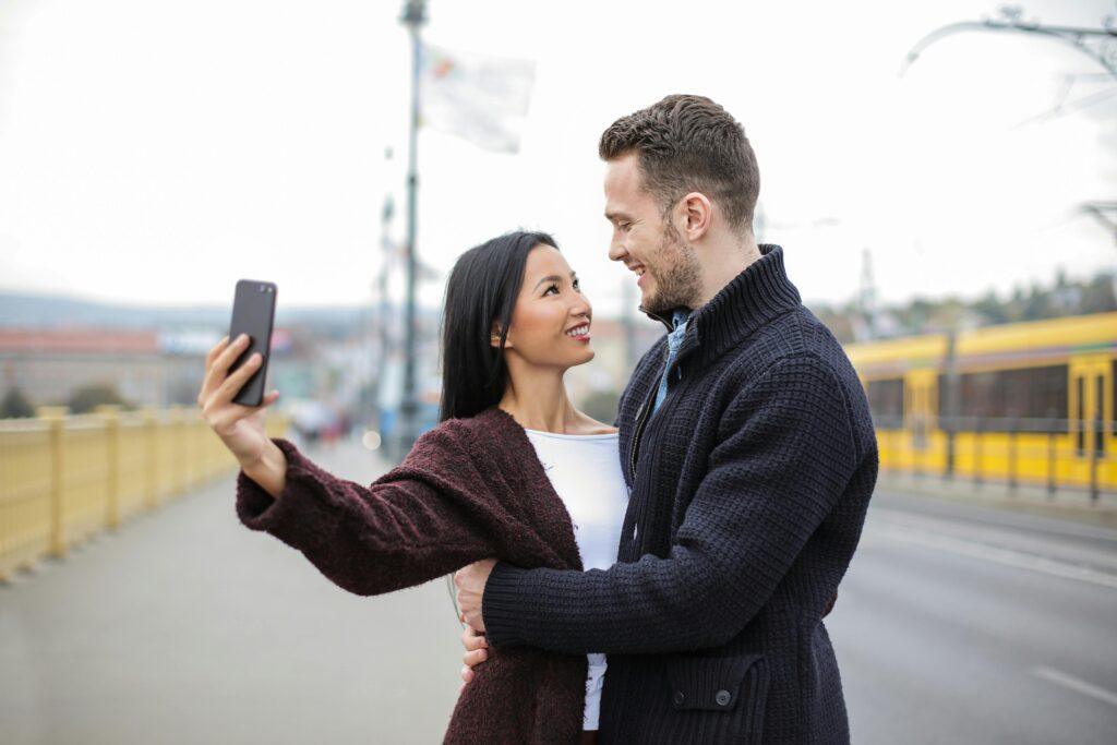A couple smiling and taking a selfie together on a city street, with a tram in the background. The woman holds the phone while they both look at each other lovingly, capturing a sweet and intimate moment perfect for a date night post with a flirty caption.