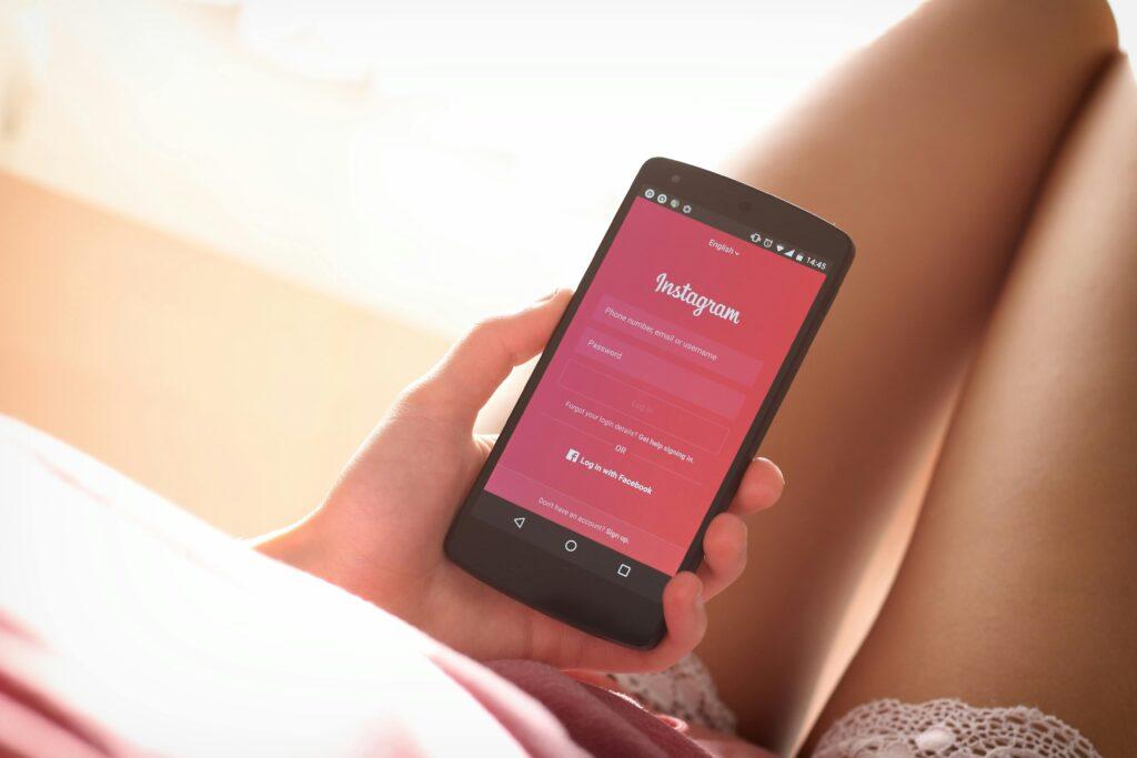 A woman relaxing with a smartphone, displaying the Instagram login screen, in her hand. The soft, natural light and cozy setting add to the inviting and relaxed atmosphere, perfect for brainstorming flirty captions for Instagram.