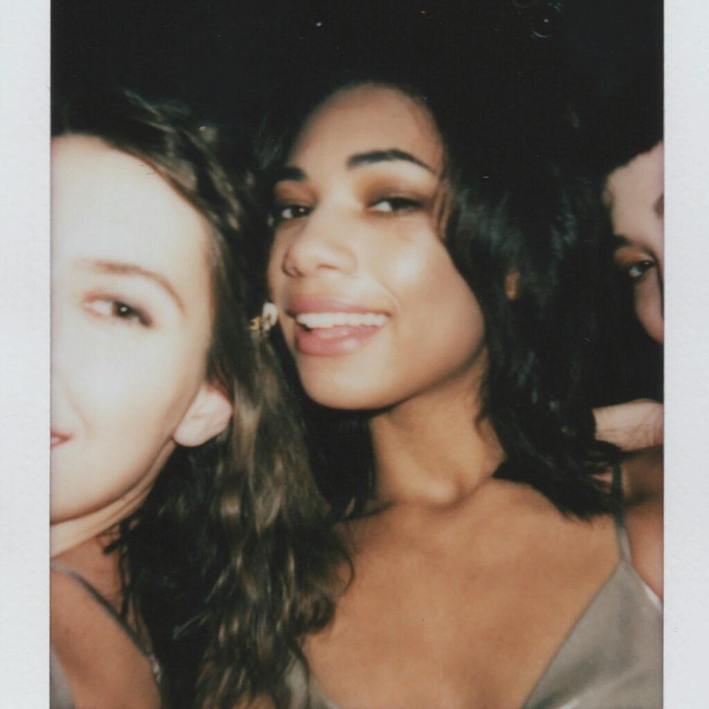 A candid polaroid photo of three female friends, smiling and posing closely together at a party. The central figure playfully sticks out her tongue, adding a fun and flirty vibe to the moment.