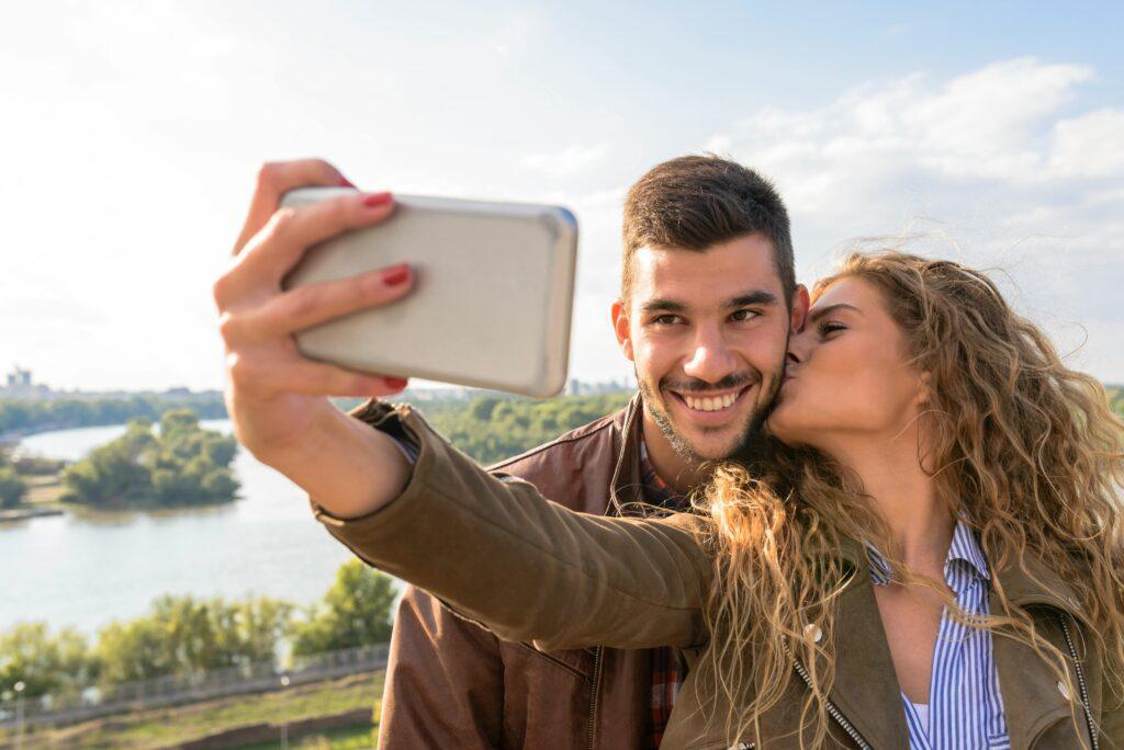 A couple taking a selfie outdoors with a scenic background, as the woman kisses the man on the cheek. Both are smiling, creating a joyful and romantic moment perfect for sharing with a flirty caption.