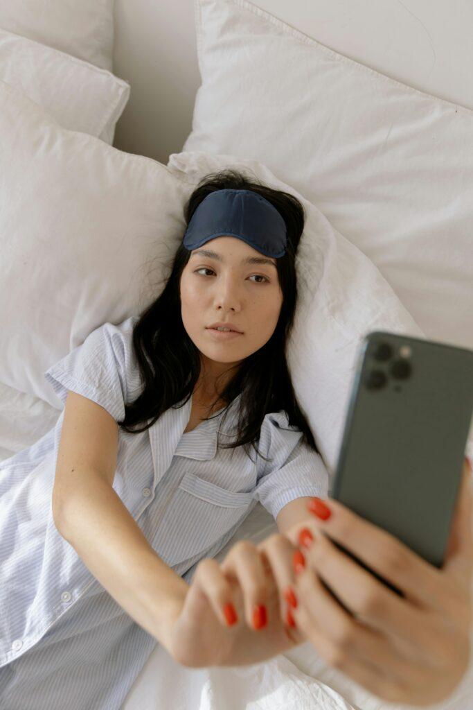 A woman lying in bed wearing a striped pajama top and a navy sleep mask pushed up on her forehead, taking a selfie with her smartphone. Her relaxed expression and cozy surroundings suggest a morning of self-care and relaxation.